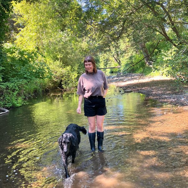 Woman walking in a shallow creek with a black dog.