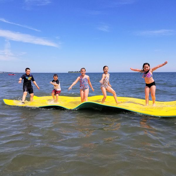 Five children playing and jumping on a floating yellow mat in the water under a clear blue sky.