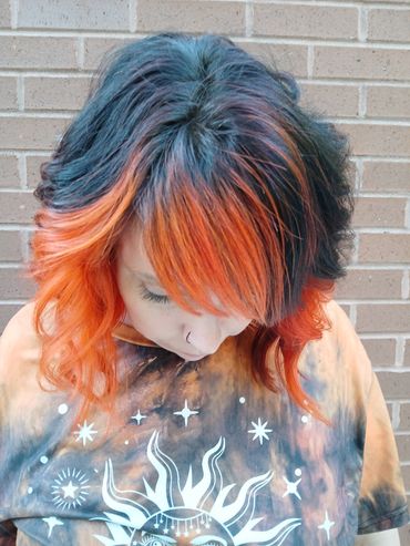 Person with black and vibrant orange hair looking down, wearing a celestial-themed tie-dye shirt.