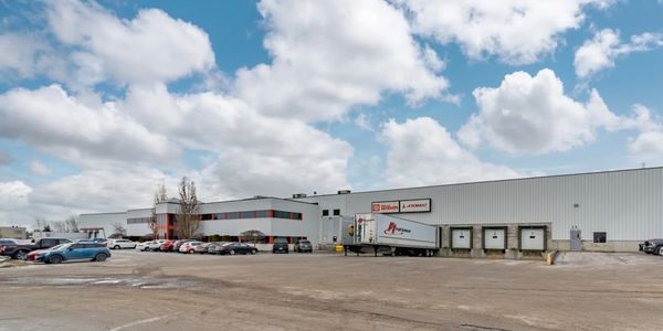 Large industrial warehouse with parked cars and loading docks under a cloudy sky.