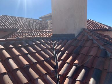 Sunlit terracotta roof tiles around a chimney under a clear blue sky.