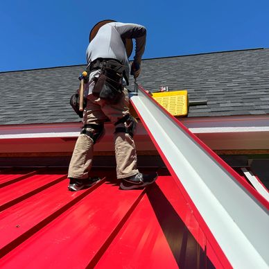 Worker installing a gutter on a red metal roof under clear blue sky.
