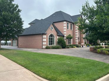 Large brick house with dark roof surrounded by greenery and trees.