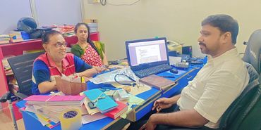 Three people engaged in a discussion at a cluttered office desk with a computer.