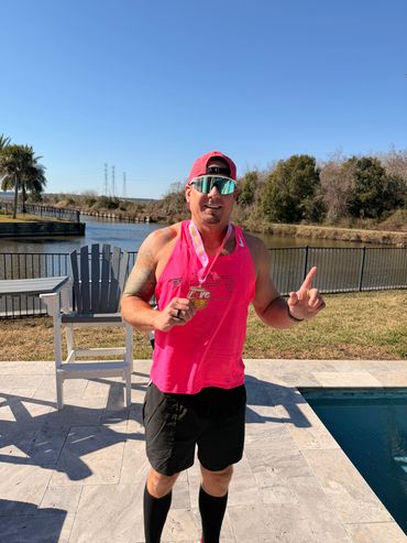 Man in pink tank top holding a medal by a pool and river.