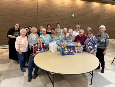 Group of elderly women posing around a table with gift baskets in a community hall.