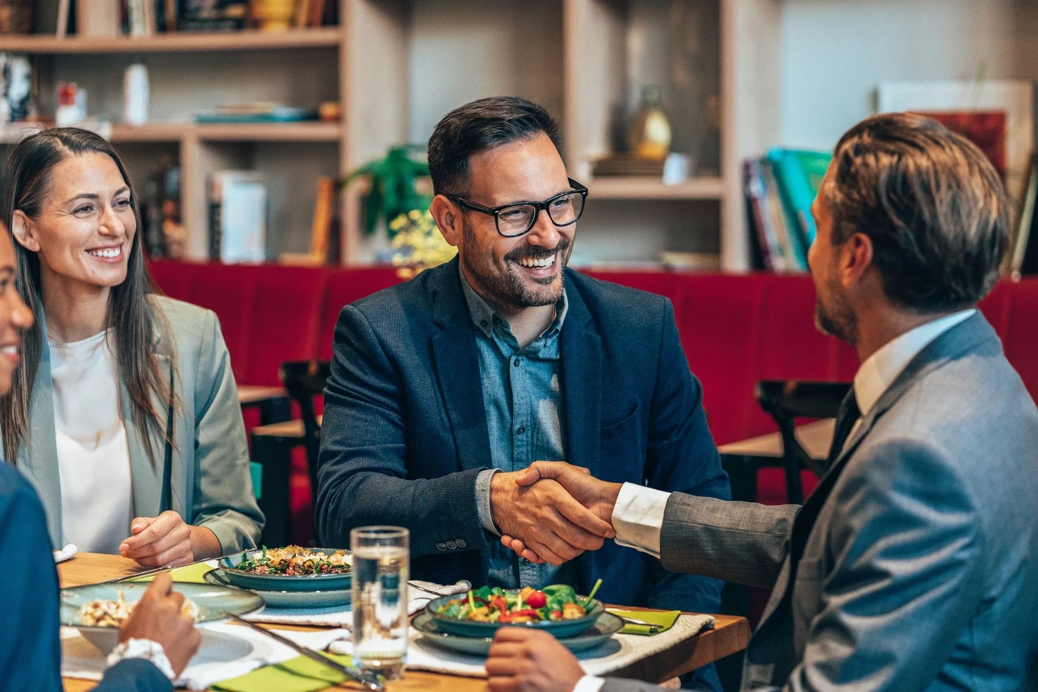 Two men shaking hands across a table during a business lunch meeting.