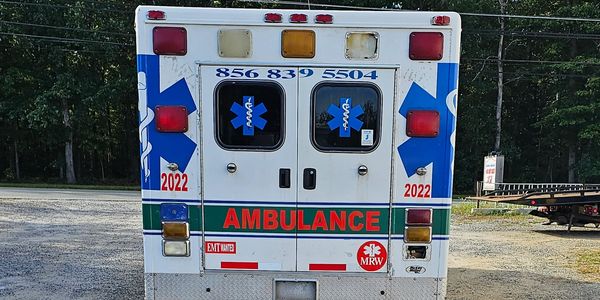 Rear view of an ambulance parked on a gravel lot with trees in the background.