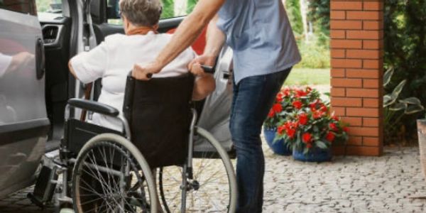 A man helps an elderly person in a wheelchair into a vehicle.