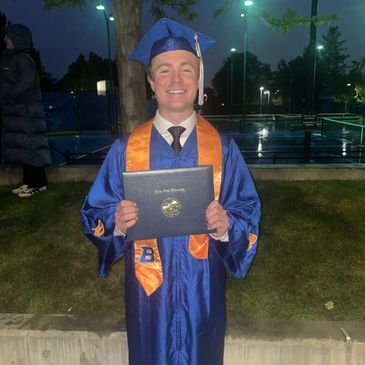 Smiling graduate in blue cap and gown holding diploma outdoors at night.