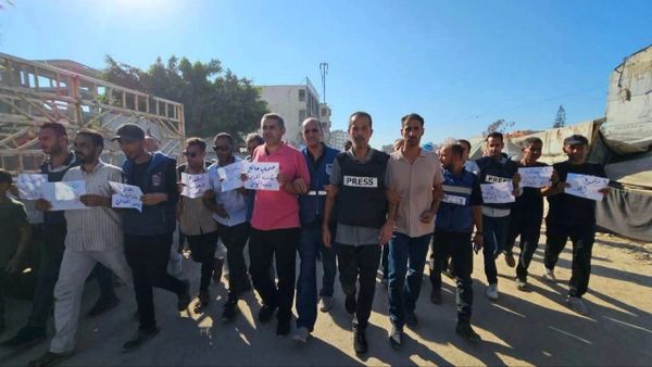 Group of men, some wearing press vests, holding signs and marching together under a clear blue sky.