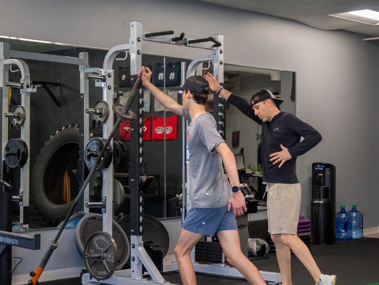 A trainer assists a young man with a weighted exercise in a gym.