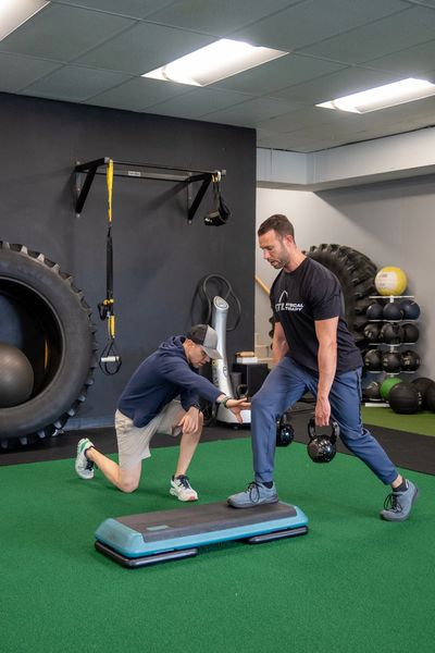 Trainer assists a man doing lunges with kettlebells in a gym.