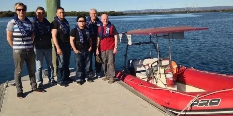 Six men wearing life jackets stand on a dock next to a red motorboat on a sunny day.