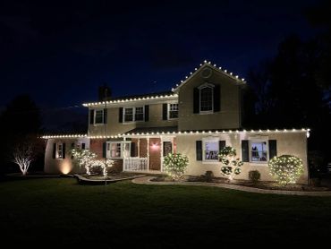A house decorated with white holiday lights along the roof and bushes at night.