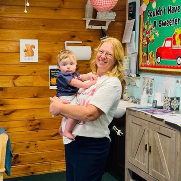 Woman smiling while holding a baby in a cozy classroom.