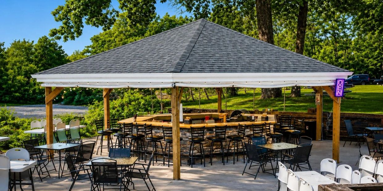 Outdoor bar with seating under a pavilion surrounded by trees.