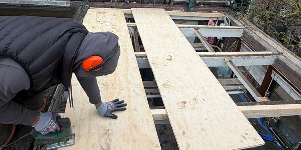 Worker installing plywood sheets on a roof with safety gear.