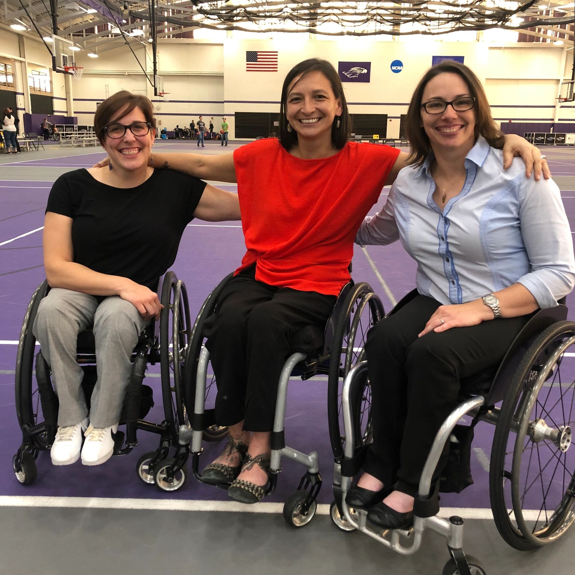 Three women in wheelchairs with arms on each others' shoulders and smiling set in a gymnasium