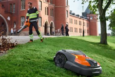 A robotic lawn mower operates on a grassy hill near a worker with a leaf blower.