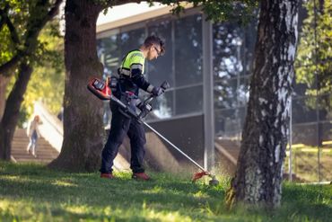 A worker trims grass with a string trimmer in an urban park.