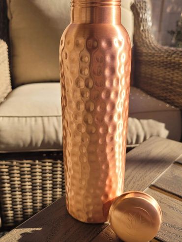 A hammered copper water bottle with its lid placed beside it on a wooden table.