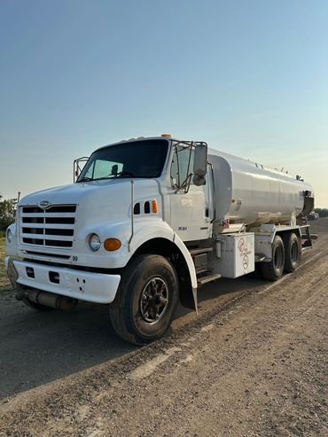 White tanker truck parked on a dirt road under clear blue sky.