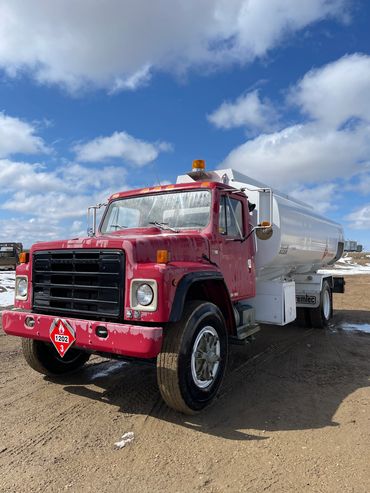 Red and white fuel tanker truck parked on a dirt road under a partly cloudy sky.