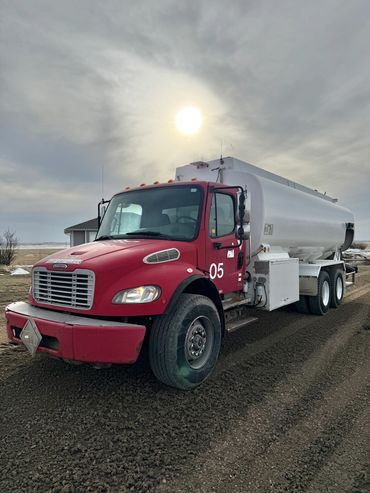 Red tanker truck parked on a dirt road under a cloudy sky with the sun shining.