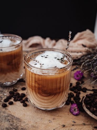 Two glasses of iced coffee garnished with herbs and flowers on a rustic wooden surface.