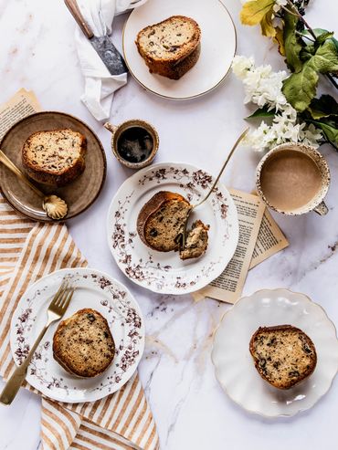 Slices of nut bread on vintage plates with coffee and flowers on a marble table.
