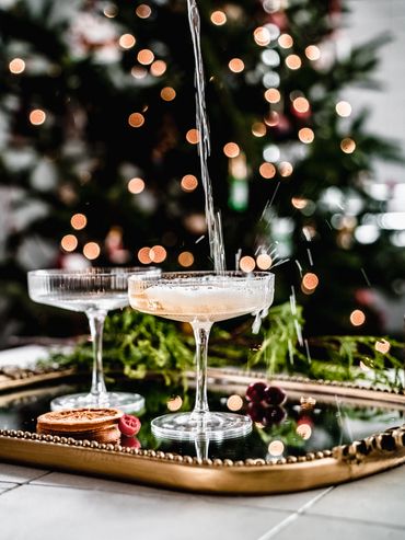 Pouring a festive drink into a coupe glass with a decorated Christmas tree in the background.