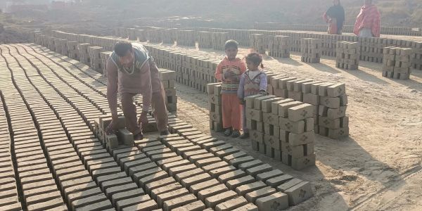 Man and children amidst rows of drying bricks in a dusty outdoor setting.
