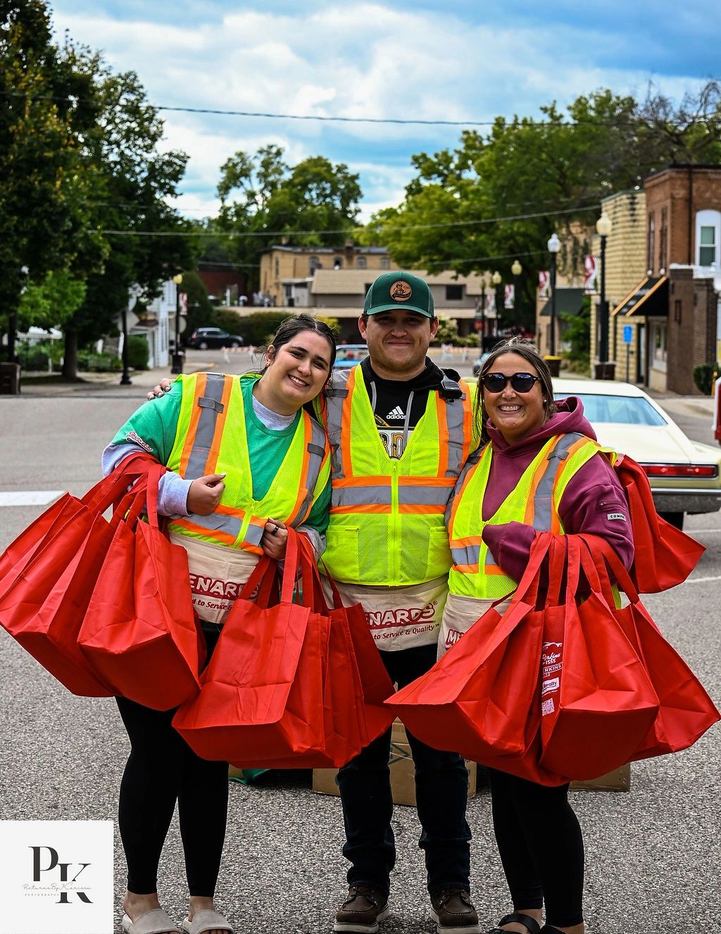 Three volunteers in safety vests holding red bags on a street.