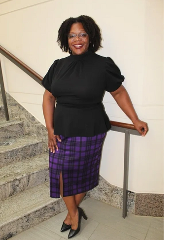 Woman in a black top and purple checkered skirt smiling on stairs.