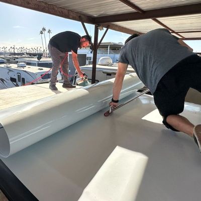 Two men installing a large white vinyl sheet on an RV roof under a metal canopy.