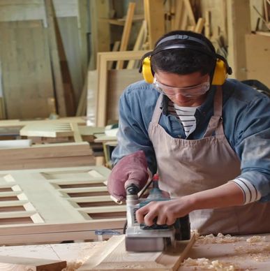 A woodworker wearing safety gear planes a wooden board in a workshop.