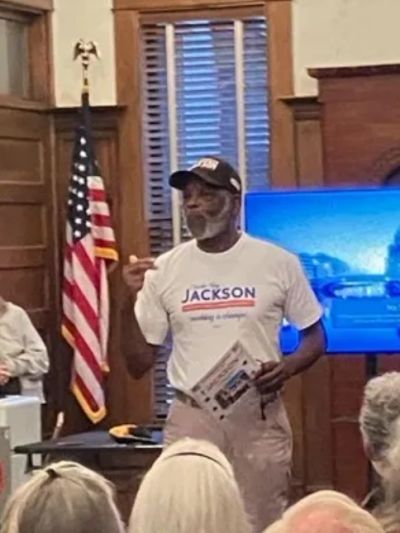Man wearing a 'Jackson' campaign shirt speaking indoors with an American flag behind him.