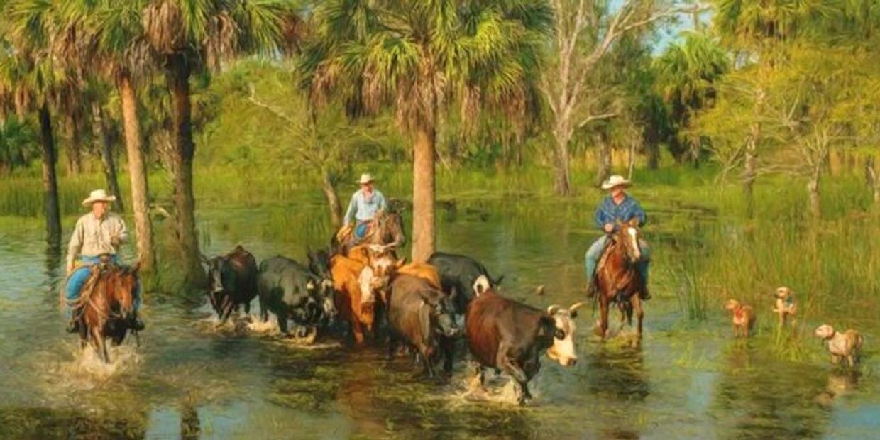Cowboys on horseback herding cattle through a shallow water area with palm trees.