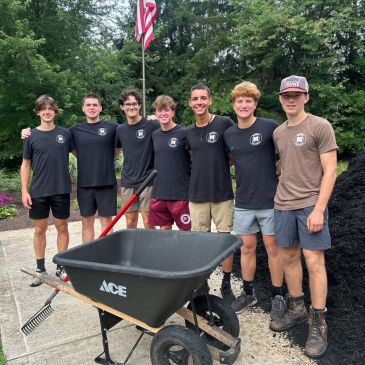 Group of seven teenagers posing near a wheelbarrow.