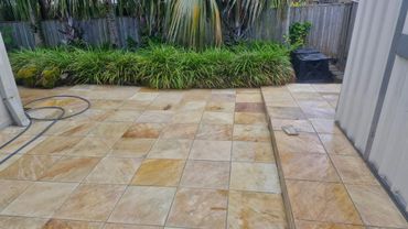 Tiled patio with lush greenery along a wooden fence and a garden hose coiled on the ground.