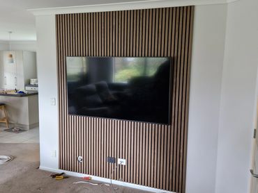 Modern TV mounted on a wooden slat wall panel with tools scattered on carpet.