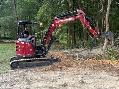 A man operating a red excavator in a wooded area.