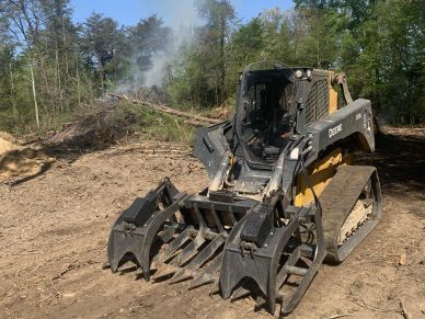 John Deere 333G loader at a clearing site with smoke in the background.