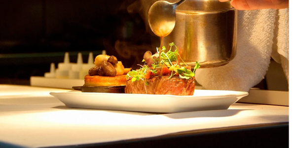Chef pouring sauce over a gourmet steak garnished with microgreens on a white plate.