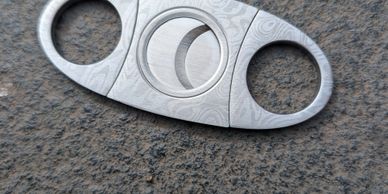 Sleek silver cigar cutter with intricate pattern on a textured surface.