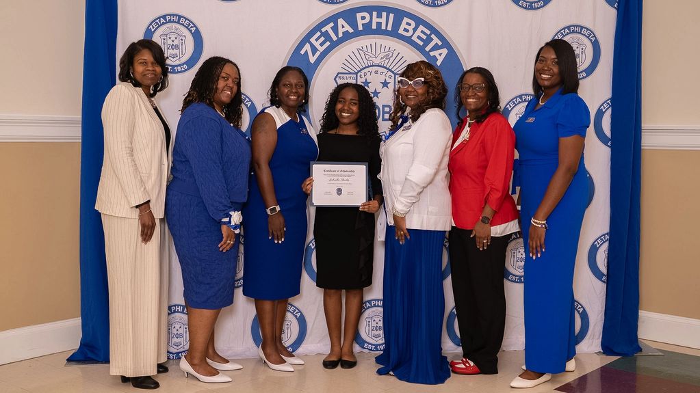 Seven women pose in front of a Zeta Phi Beta banner, one holding a certificate.