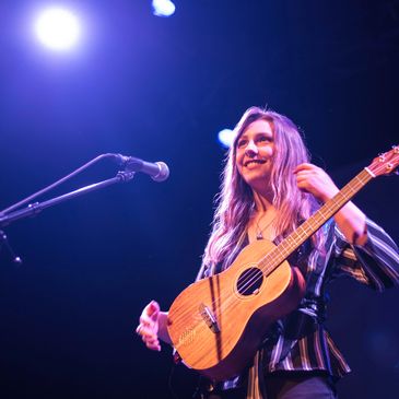 Larissa standing on stage with a microphone and a baritone ukulele