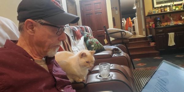 Man with glasses and cap sitting on couch with cream-colored cat beside him.