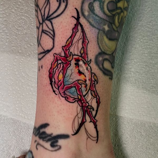 Colorful tattoo of a cracked egg with red claw-like structures on a person's ankle.